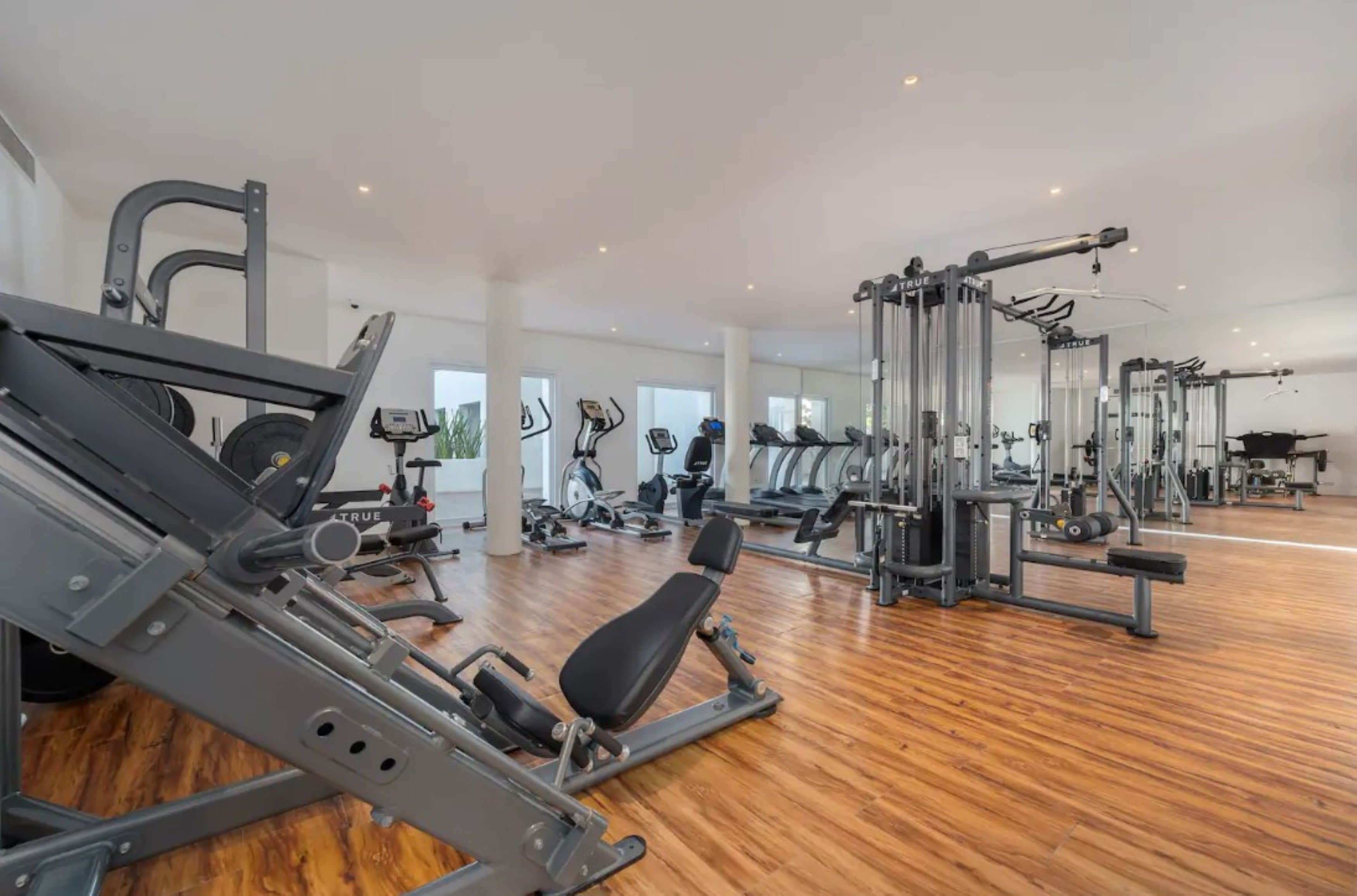 Une salle de sport spacieuse, dotée de divers appareils d’exercice, de grandes fenêtres et d’un plancher en bois.