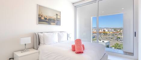 A modern hotel room with a large bed, white bedding, a nightstand with a lamp, a framed cityscape picture, and a view of a cityscape through a large window.