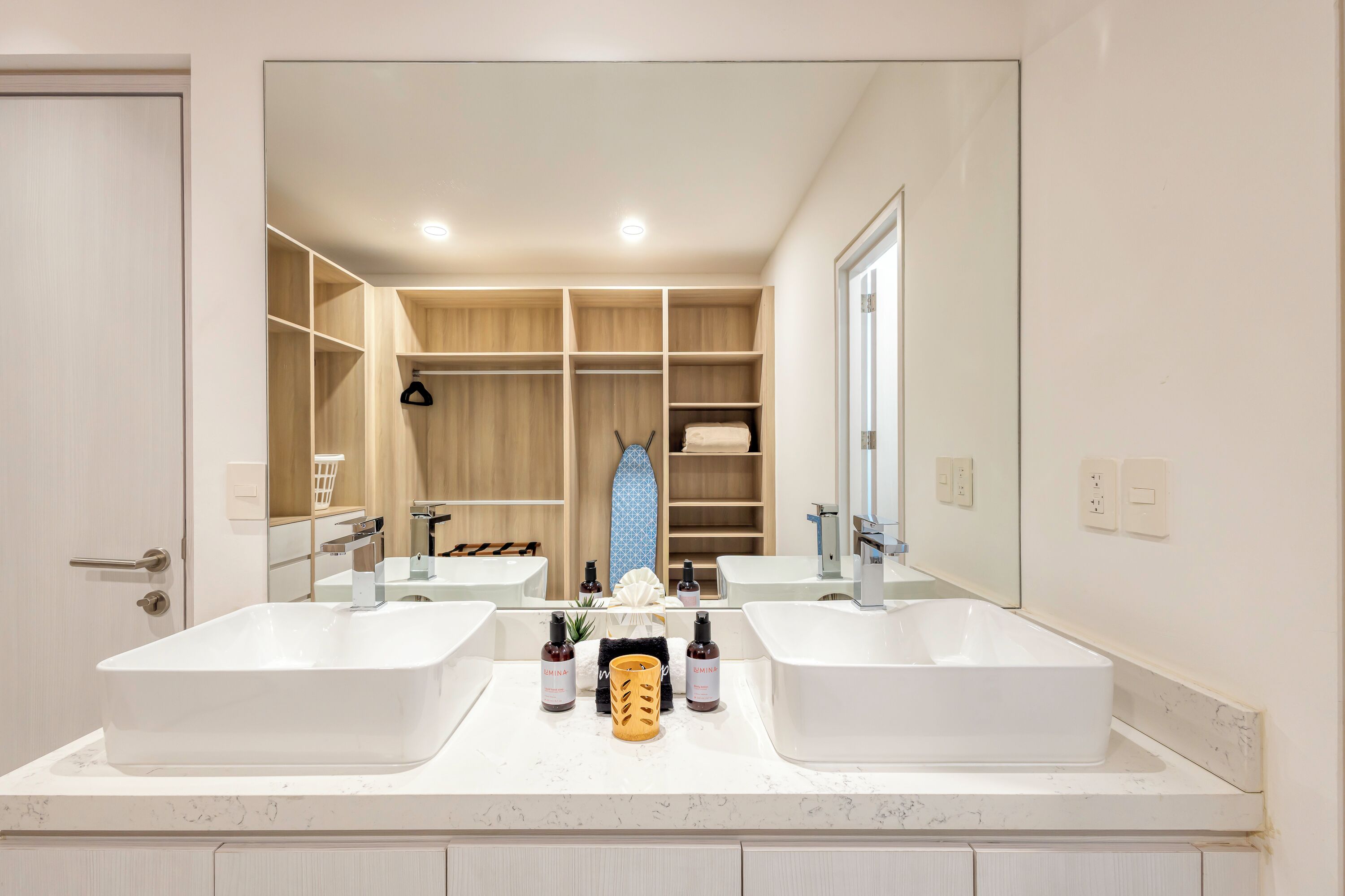 A modern bathroom with two sinks, a large mirror, and a walk-in closet with wooden shelving.