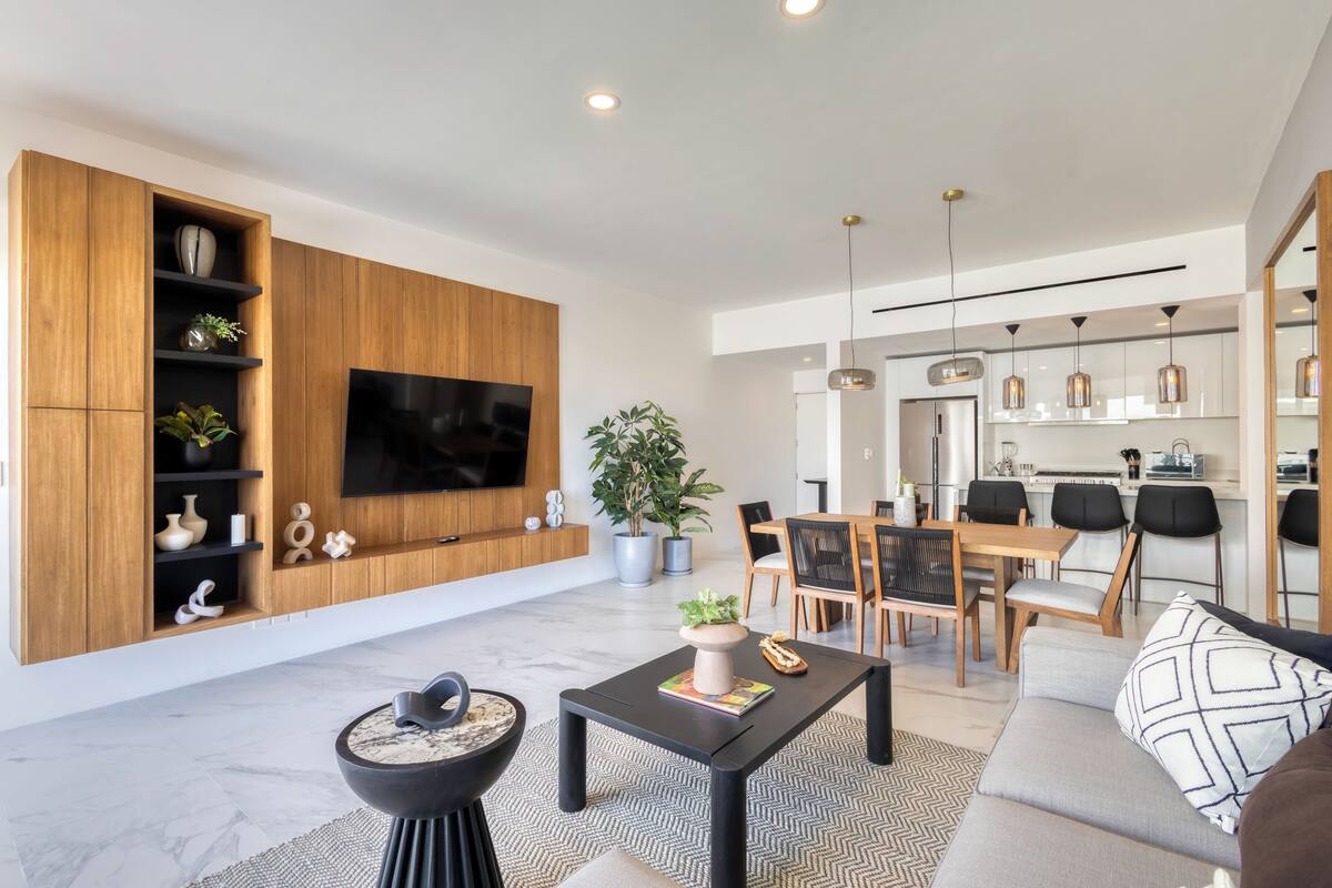Una sala moderna con televisor de pantalla plana, estanterías de madera, un comedor con mesa y sillas, y una cocina al fondo.