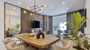 A modern living room with a wooden dining table, beige chairs, a large potted plant, and a flat-screen TV mounted on the wall.
