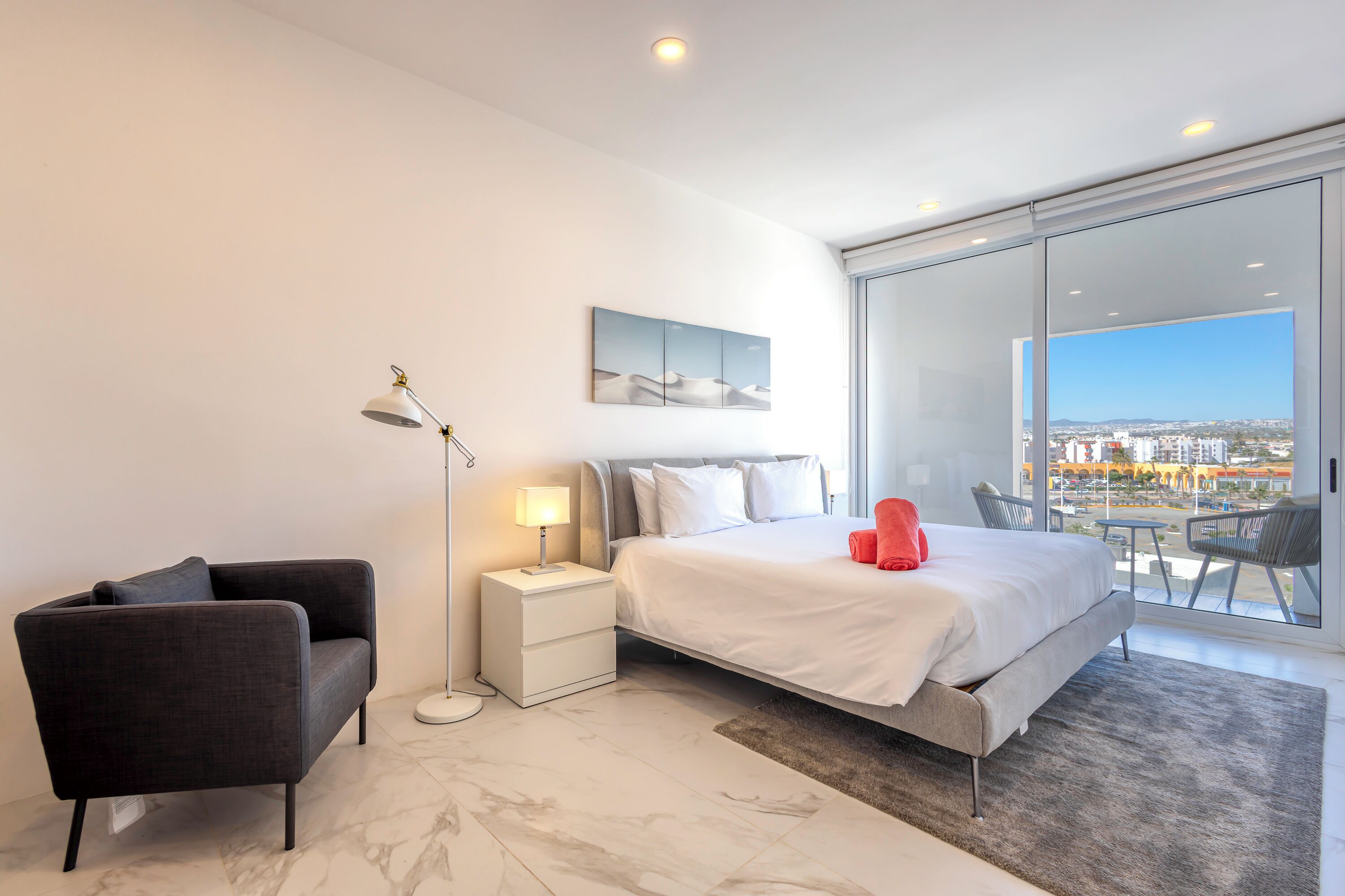 Une chambre moderne avec un grand lit, une table de chevet, une chaise et une vue sur la ville grâce à de grandes fenêtres.