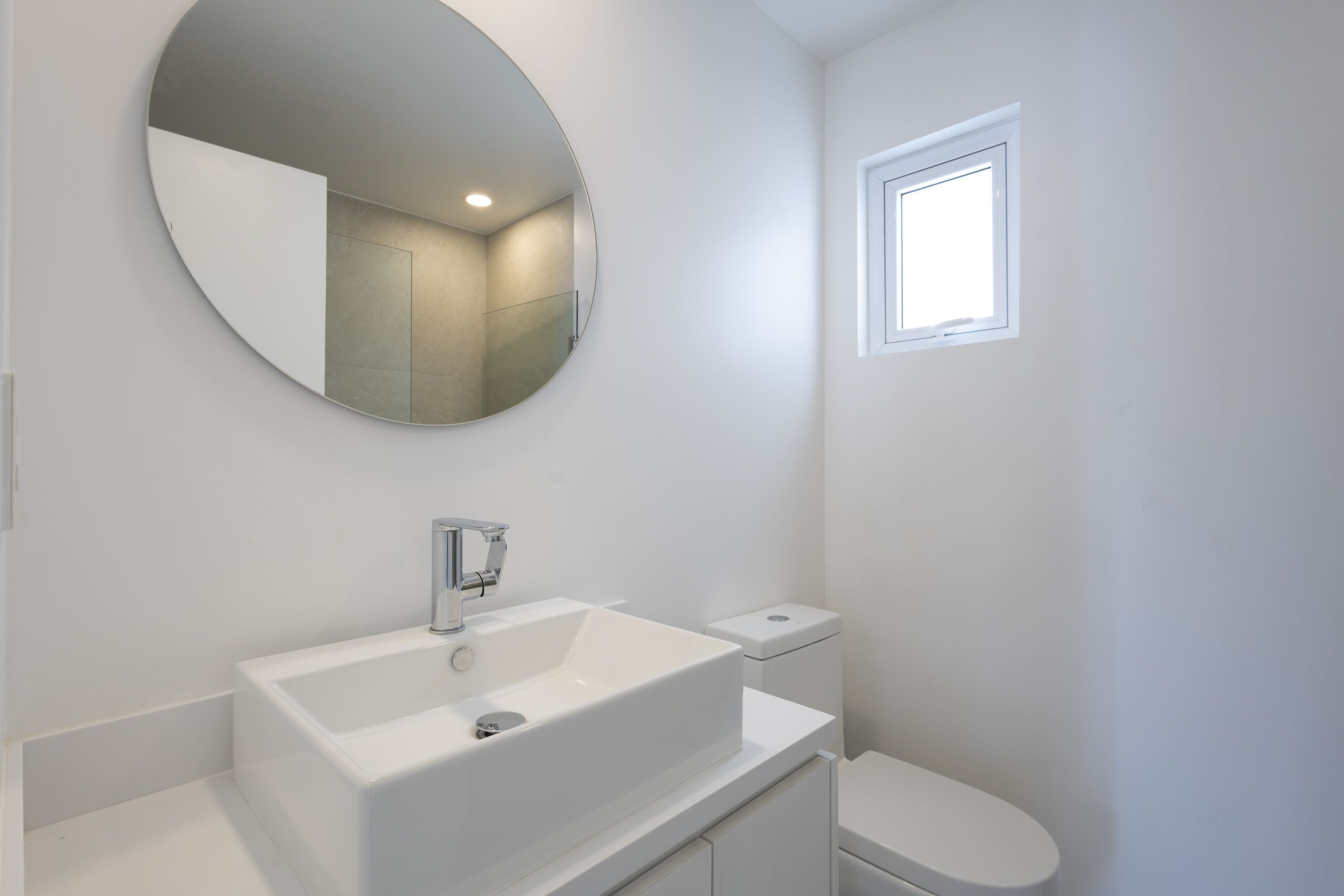 A modern bathroom with a white sink, a round mirror, and a window.