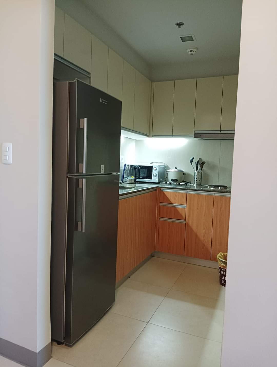 A compact kitchen with a stainless steel refrigerator, a microwave, and a toaster. The cabinets are light wood, and there is a trash bin in the corner.