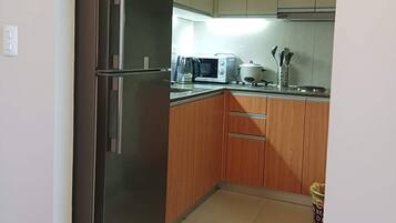 A compact kitchen with a stainless steel refrigerator, a microwave, and a toaster. The cabinets are light wood, and there is a trash bin in the corner.