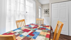 A dining area with a table covered by a colorful quilt, wooden chairs, a white door, and a window with sheer curtains. - Riverview beatiful acre home Pet/RV/boat Friendly (Riverview)