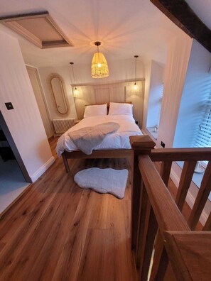 A bedroom with a wooden floor, a bed with white linens, a hanging light fixture, and a view of the outside. - Charming 1-bed Cottage in Brynmenyn (Bridgend)