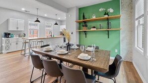 Une cuisine moderne avec des meubles blancs, une table à manger en bois pour six personnes et un mur d’accent vert.