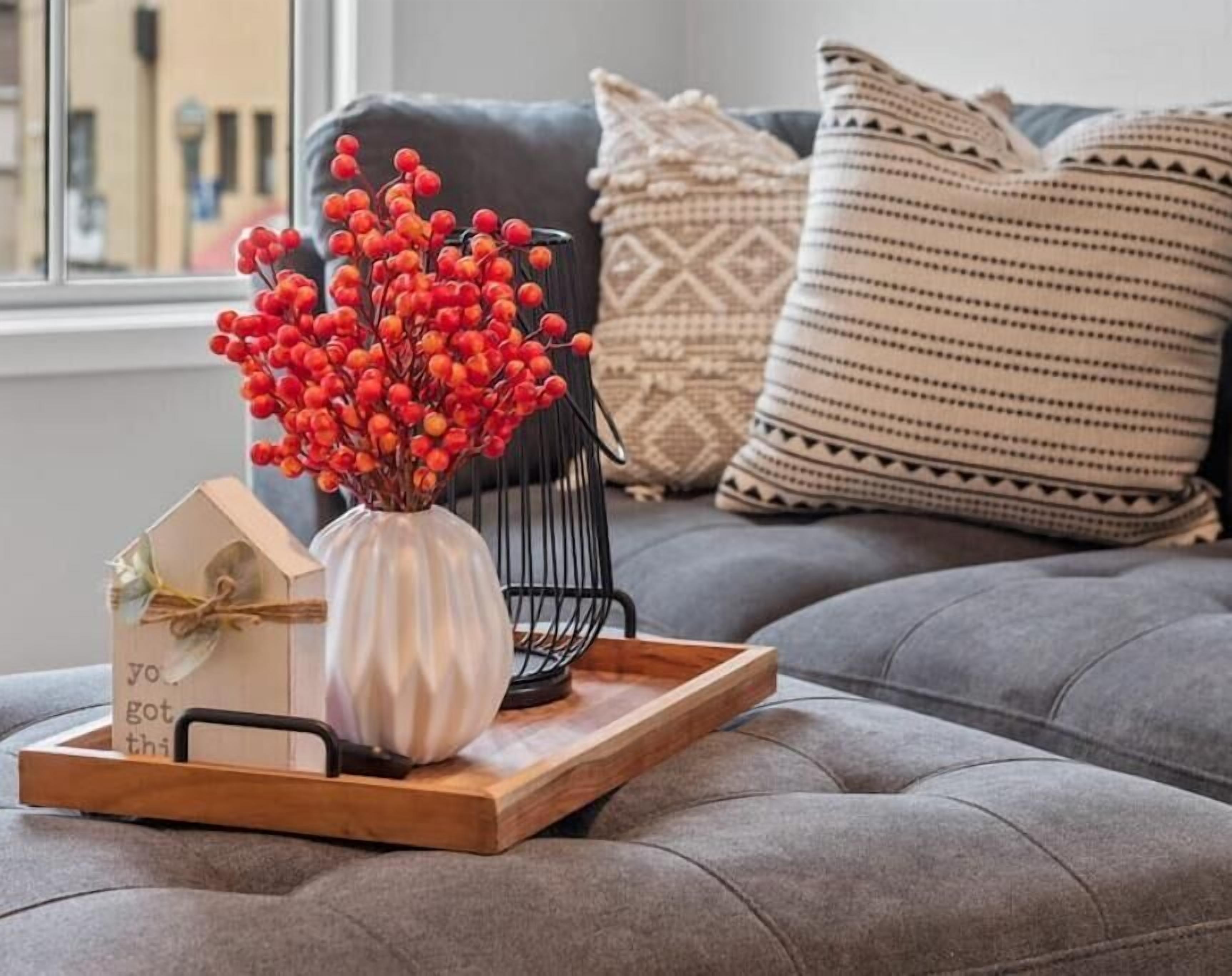 Ein gemütliches Wohnzimmer mit einem grauen Sofa, einer dekorativen Vase mit roten Beeren und einer Holztablett mit einer kartenförmigen Zeichnung eines Hauses und einer kleinen Geschenkbox.
