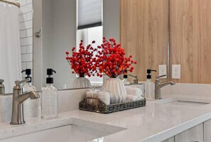 A modern bathroom with a white countertop, two sinks, and a mirror. There are decorative red berries in a vase and a basket of toiletries. - New built w/free parking off Brady St. Fiserv Forum, Summer Fest and Lakefront! (Milwaukee)