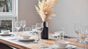 Une table à manger dressée avec de l’assiette, des bols et des verres blancs, un vase contenant des herbes des pampas séchées et une table en bois dotée d’un cadre en métal noir.