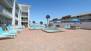 A hotel pool area with multiple lounge chairs and umbrellas, surrounded by a white building with balconies.