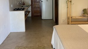 A compact kitchen with a refrigerator, a sink, and a stove. There is a wooden door leading to another room. The kitchen has a concrete floor and a ceiling with wooden beams. - Vila dos Lobatos (Porto Seguro)