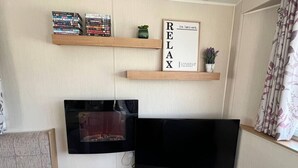Un soggiorno con TV a schermo piatto, scaffali in legno pieni di libri, un cartello con la scritta 'RELAX' e una piccola pianta.