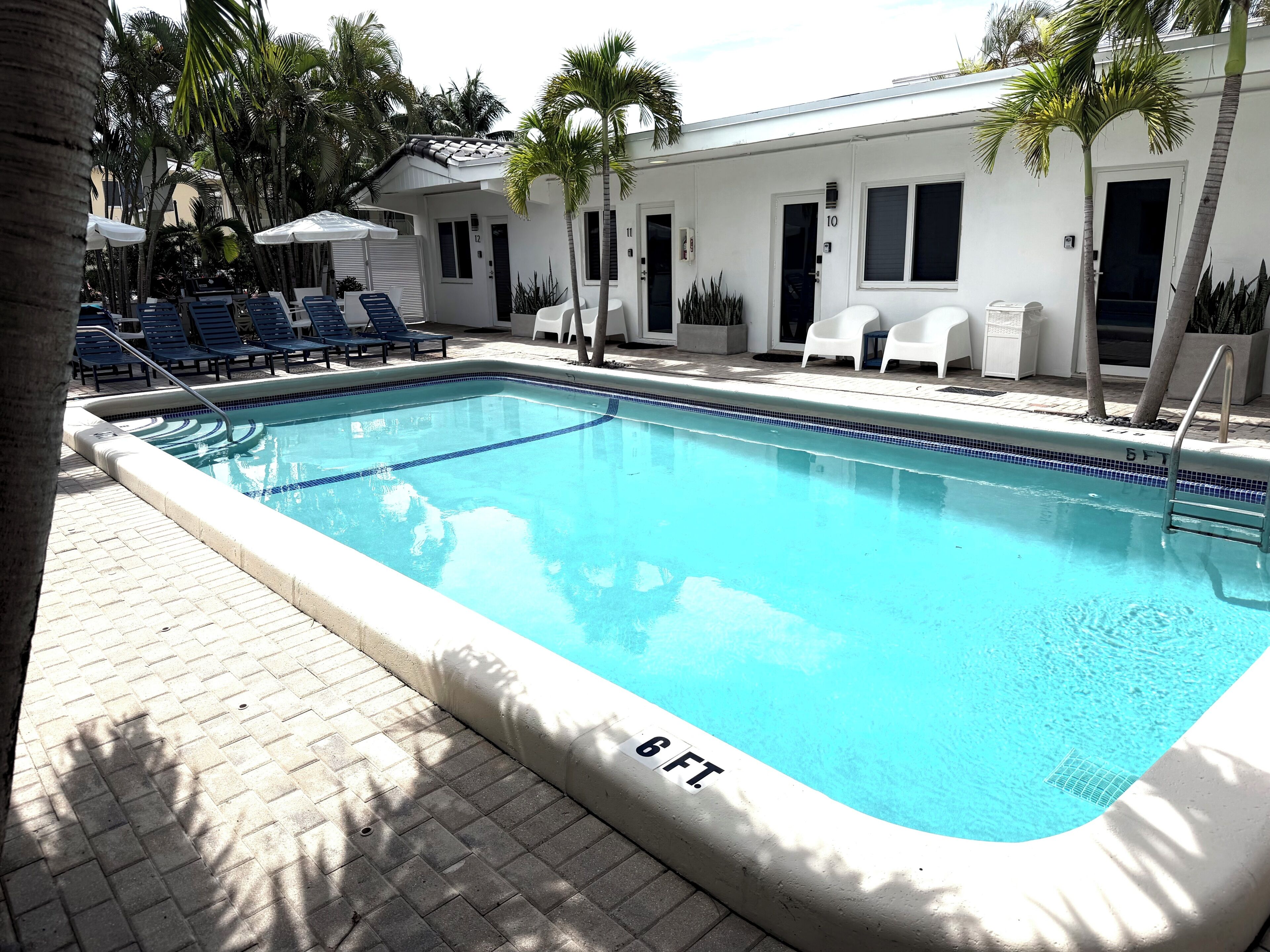 Une piscine rectangulaire dont la profondeur est indiquée à 6 pieds, entourée de chaises longues et de palmiers, avec un bâtiment visible au fond.