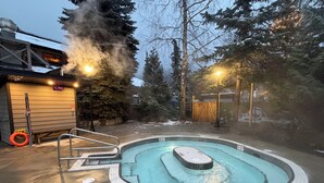 A hot tub surrounded by a wooden deck, with a sauna and a covered area in the background.