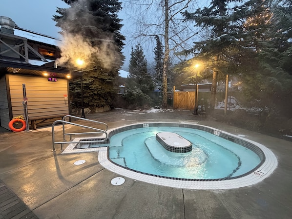 Een hottub omringd door een houten terras, met een sauna en een overdekt gedeelte op de achtergrond.