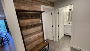 A hallway with a wooden wall, a bench, and a coat rack. There is a door leading to a bathroom with a mirror and sink.