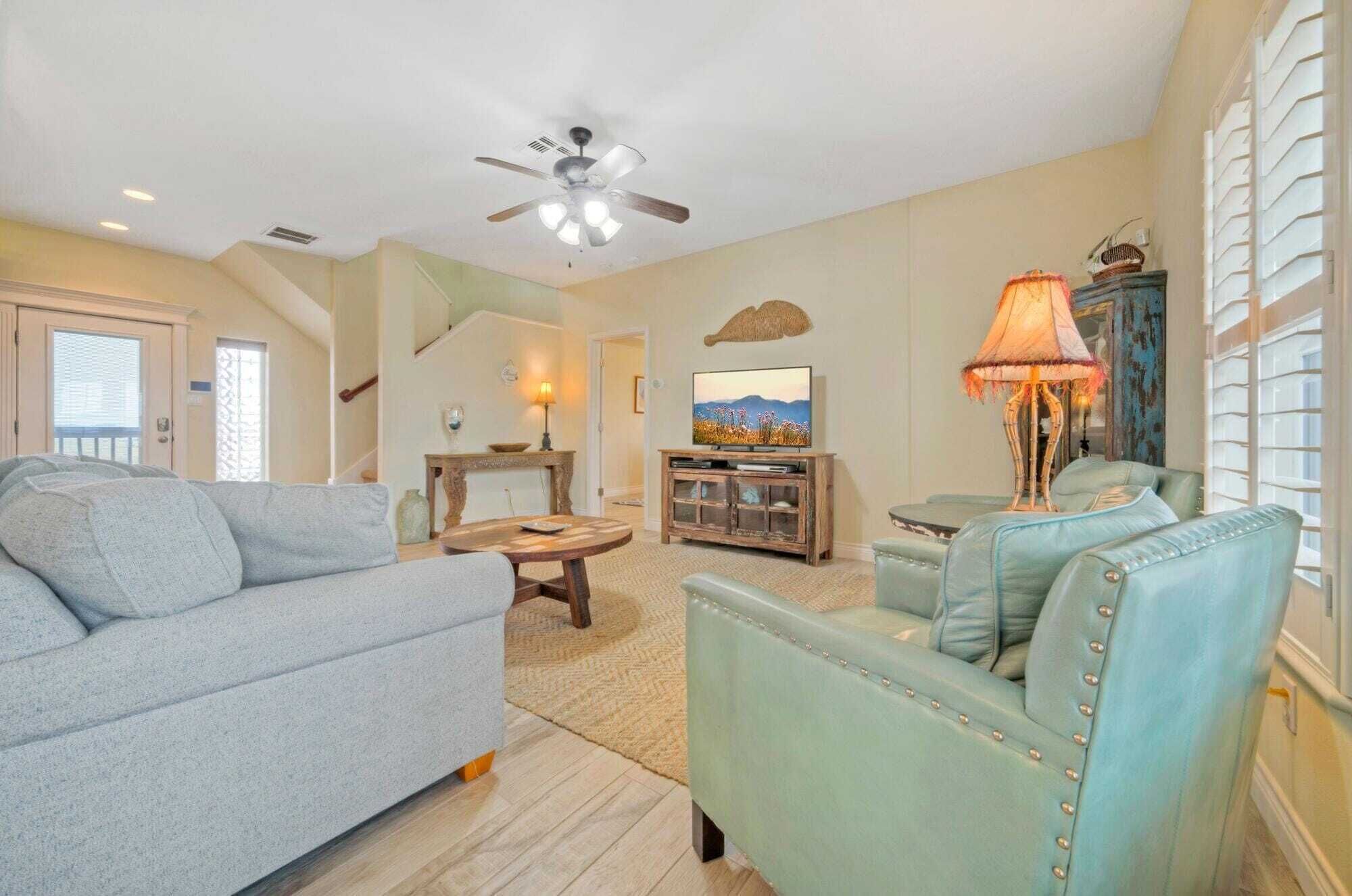 A living room with a grey sofa, a teal armchair, a wooden coffee table, and a TV on a wooden stand.
