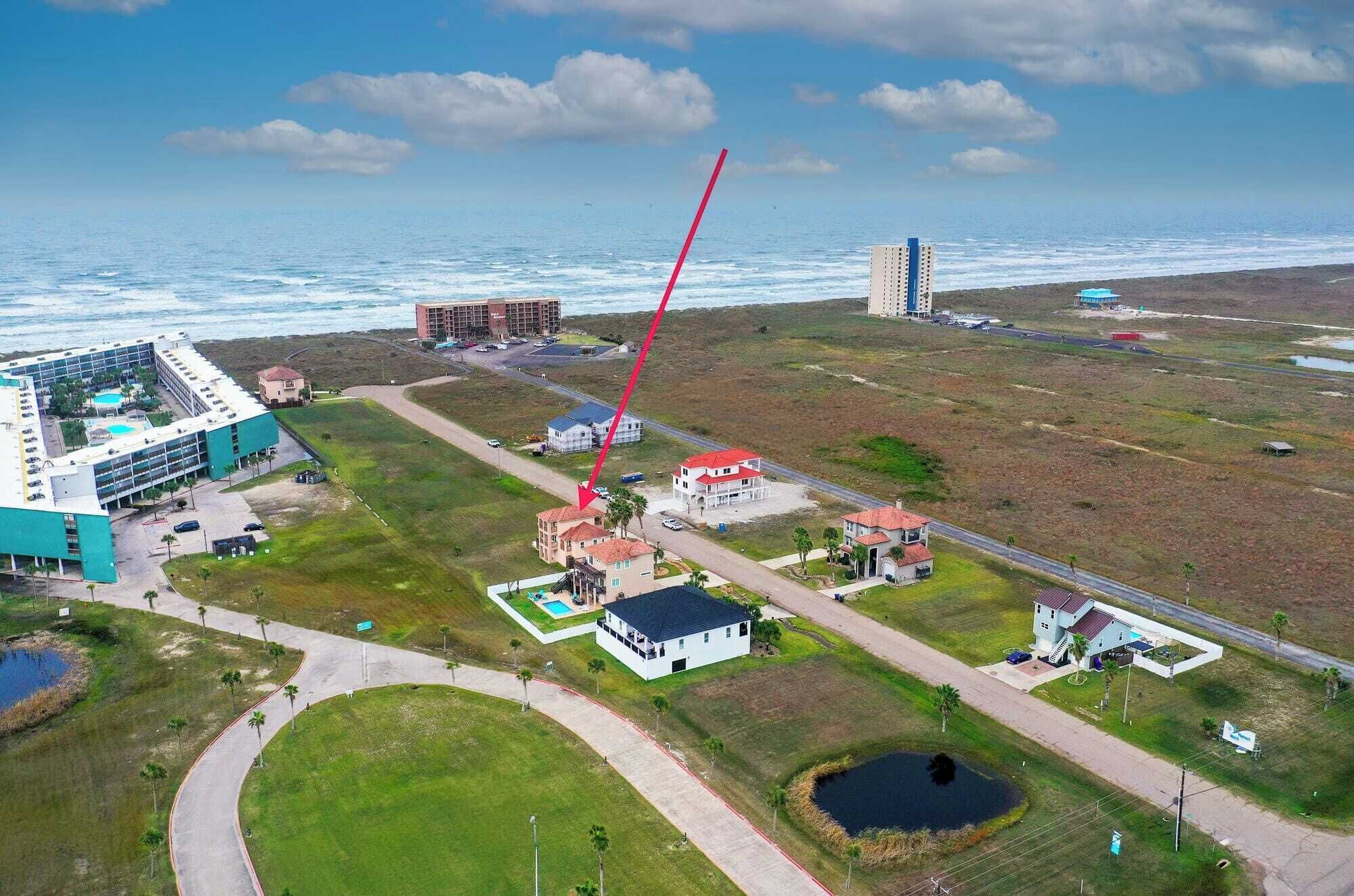 An aerial view of a coastal area with a large building complex, a road, and several residential buildings.