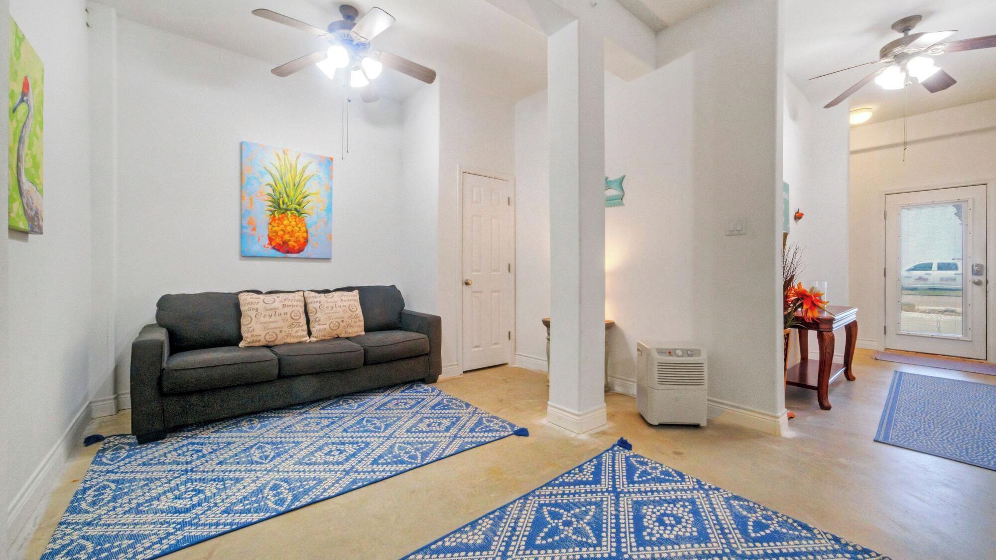 A living room with a gray sofa, blue patterned rugs, a pineapple painting, and ceiling fans.