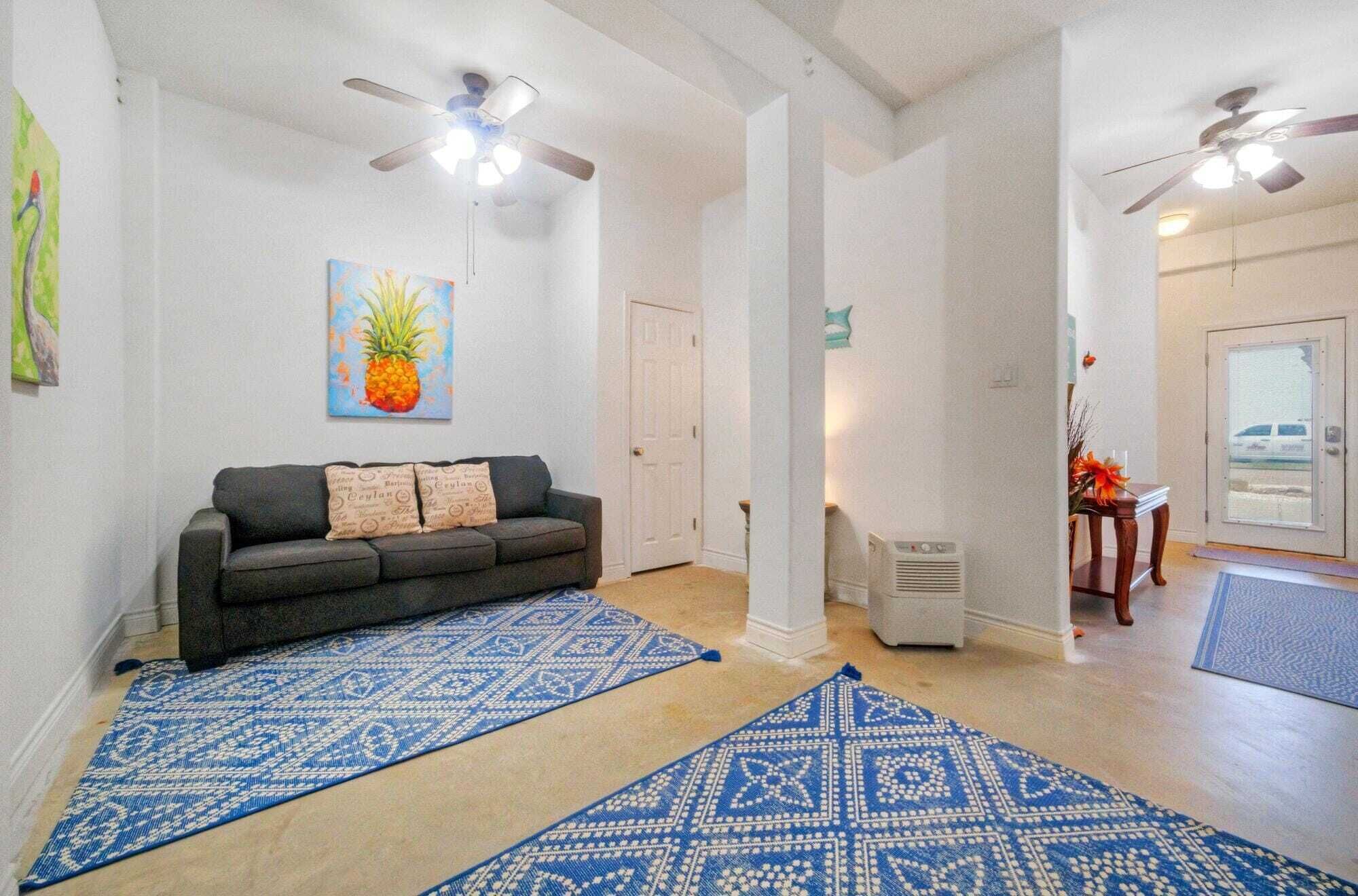 A living room with a dark-colored sofa, patterned blue rugs, a pineapple painting, and ceiling fans.