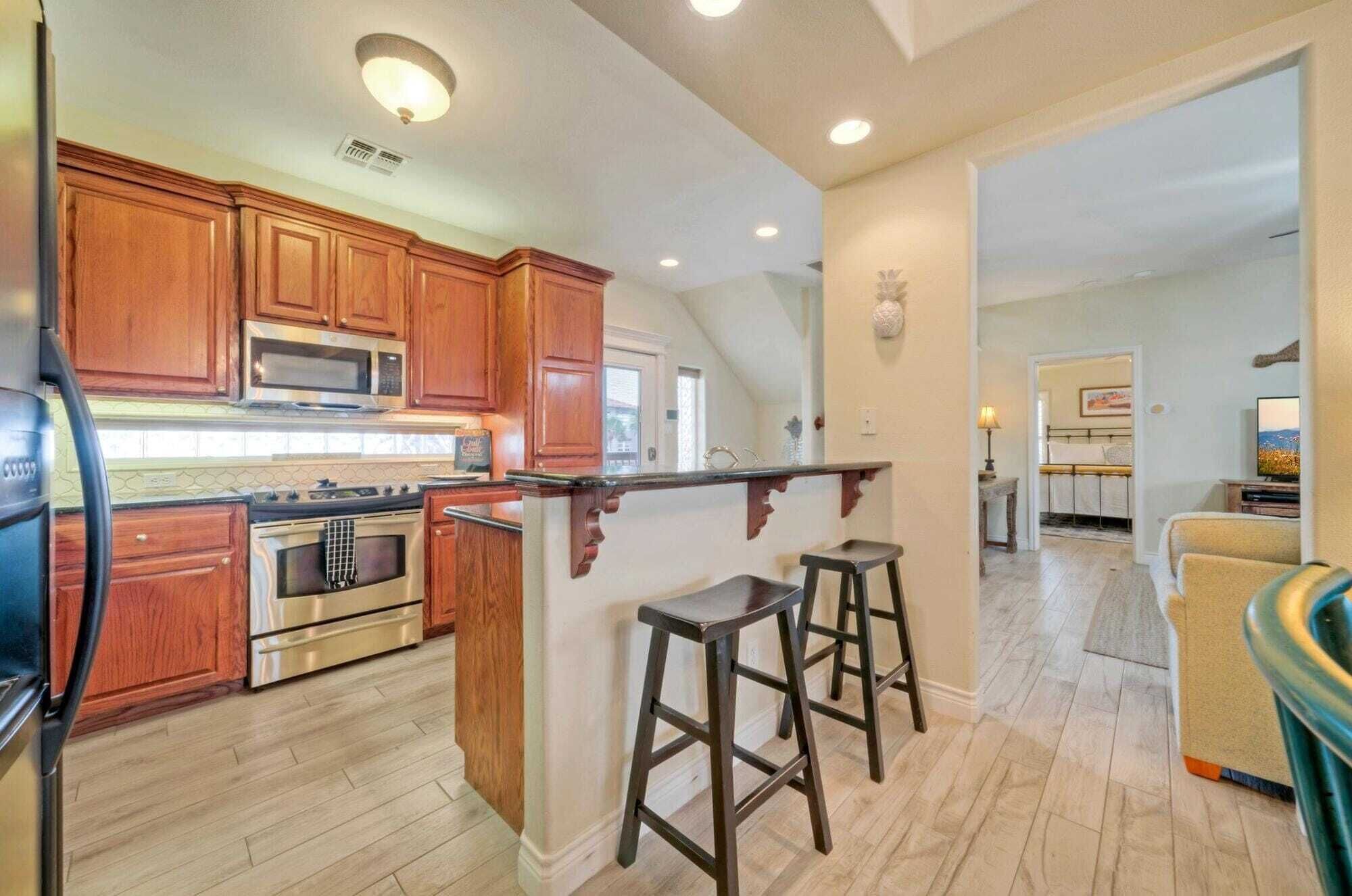 A kitchen with wooden cabinets, a microwave, and a stove. There are two stools and a bar counter.