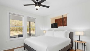 A bedroom with a large bed, two bedside tables with lamps, a ceiling fan, and a wall-mounted tapestry.