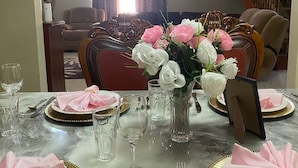 A dining table set for four with pink napkins, a vase of white and pink flowers, and a clock on the wall. - Lachemar Guest House (Ocho Rios)