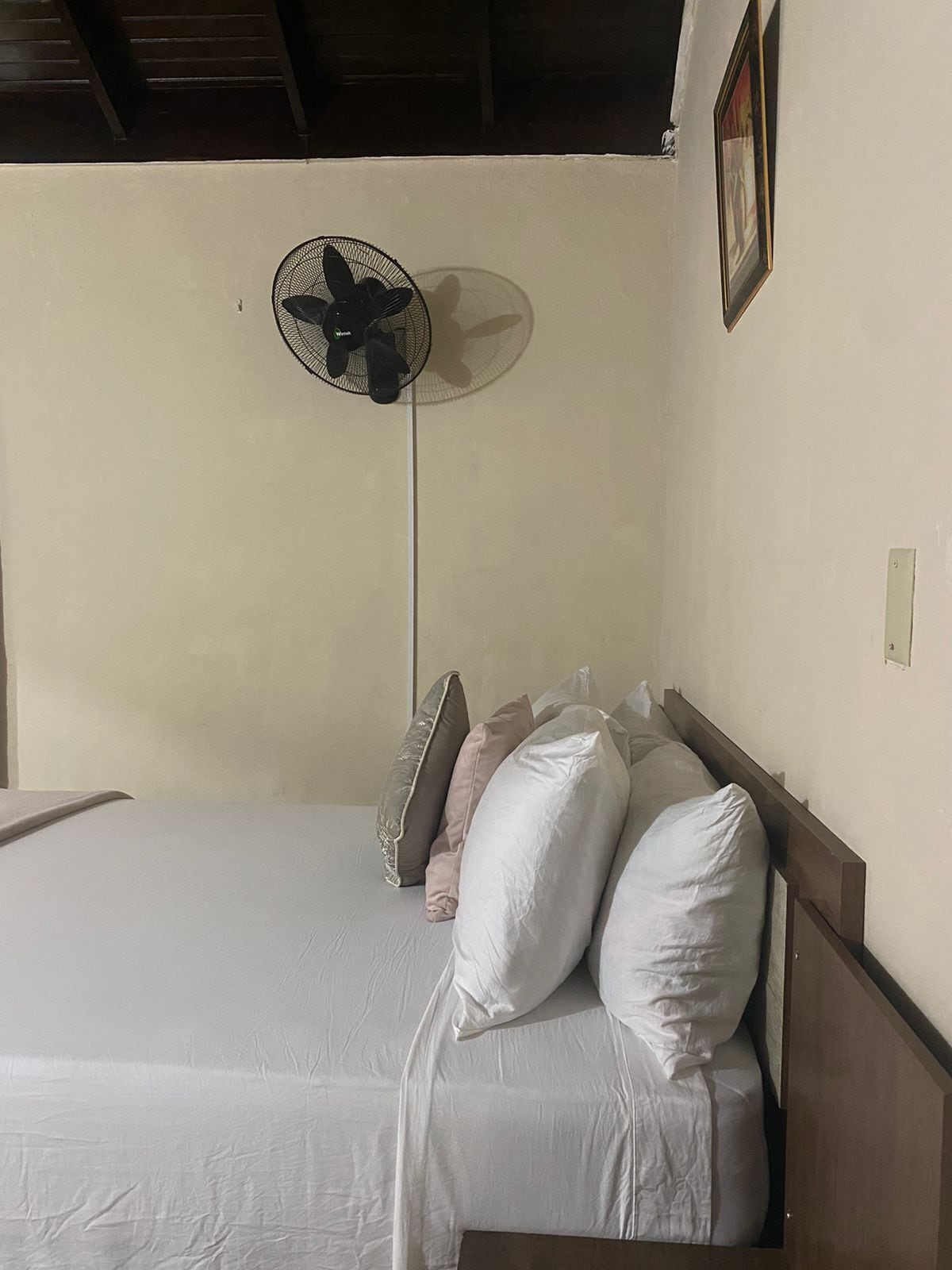 A single bed with white bedding and multiple pillows, a standing fan, and a wooden headboard.