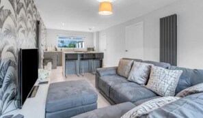 A modern living room with a grey sofa, a flat-screen TV, and a kitchen area in the background. - West Lothian 5 Beds + car space (Whitburn)