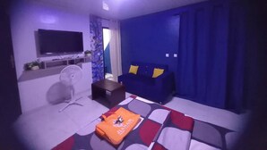 Een woonkamer met een flatscreen televisie, een bank met kussens, een salontafel en een ventilator.