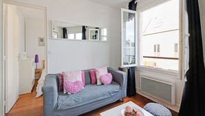 A modern living room with a grey sofa, a white coffee table, and a large window with a view of buildings. - Harbor View Hideaway (Isigny-sur-Mer)
