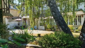 A garden with a tree, a patio, and a building with a white facade and a sloped roof. - Romantic Escape in Normandy (Isigny-sur-Mer)