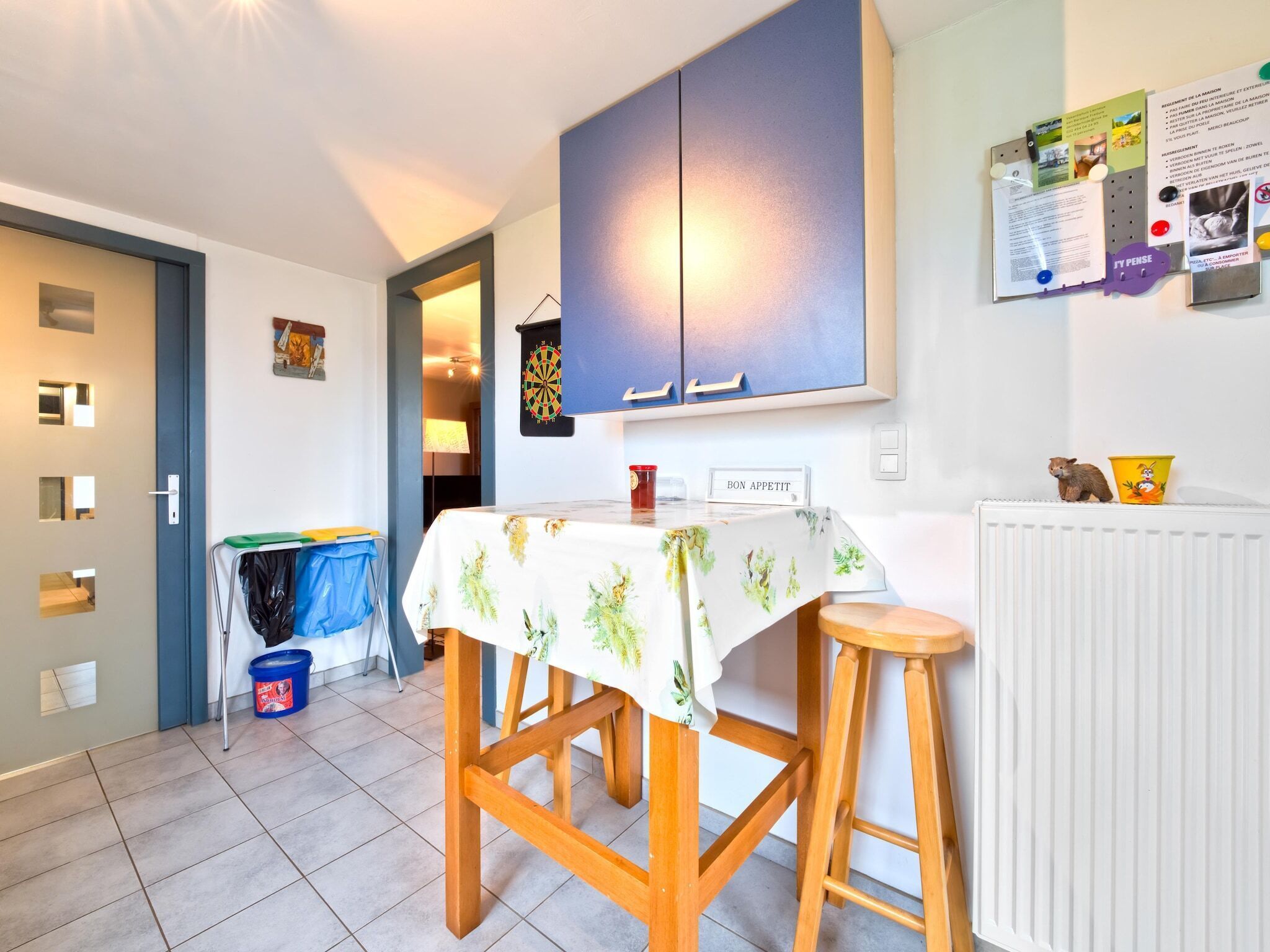 Een kleine keuken met een dartbord, een tafel met tafelkleed en een prikbord met verschillende berichtjes.