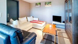 A modern living room with a white sofa, a black leather couch, a wooden coffee table, and a flat-screen TV. - Holiday Home with Garden (Lierneux)