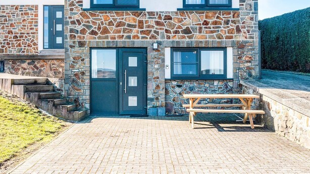 A two-story stone building with a dark blue door and windows, a paved patio with a wooden bench, and a stone wall. - Holiday Home with Garden (Lierneux)