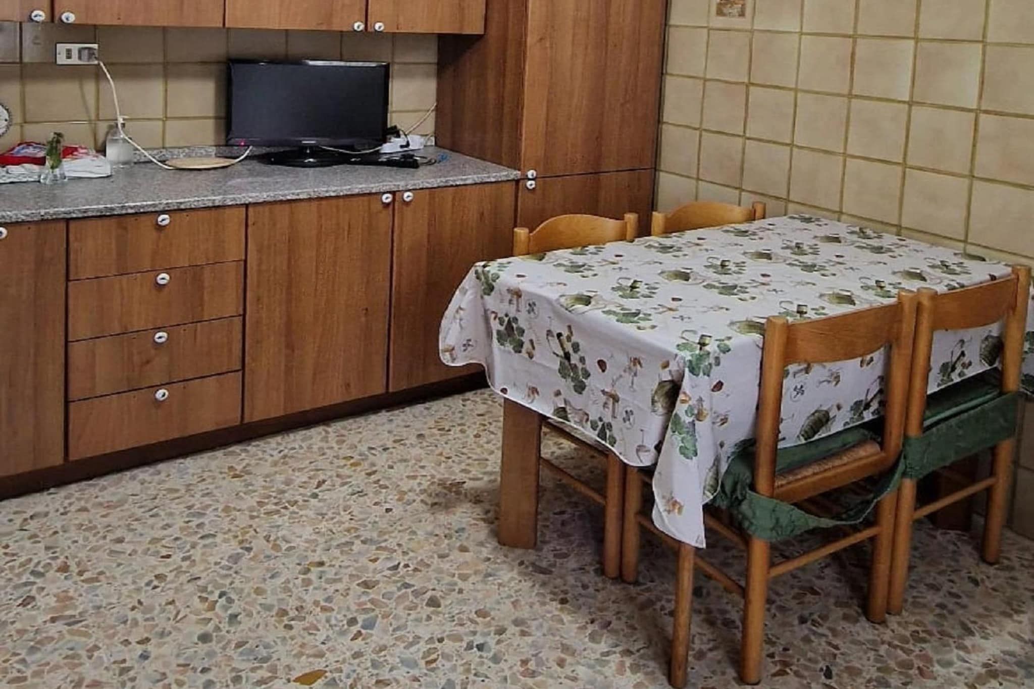 Una cucina con mobili in legno, un televisore, un tavolo con tovaglia floreale e una sedia con cuscino verde.