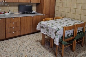 Une cuisine avec des meubles de rangement en bois, un téléviseur, une table recouverte d’une nappe à motifs floraux et une chaise dotée d’un coussin vert.