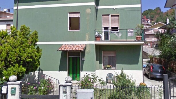 Un edificio residencial de dos plantas con fachada verde, techo de tejas y un pequeño balcón con plantas en macetas.