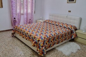 Une chambre à coucher avec un lit, une table de chevet, un tapis blanc et moelleux, et une fenêtre avec des rideaux.