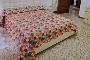 Un lit recouvert d’une couette aux couleurs vives, un radiateur et une porte dans une pièce dont le sol est carrelé.