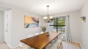 A dining area with a wooden table, white chairs, a chandelier, and a large abstract painting on the wall.