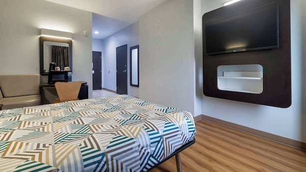 A hotel room with a bed, a desk, a chair, a TV mounted on the wall, and a mirror.
