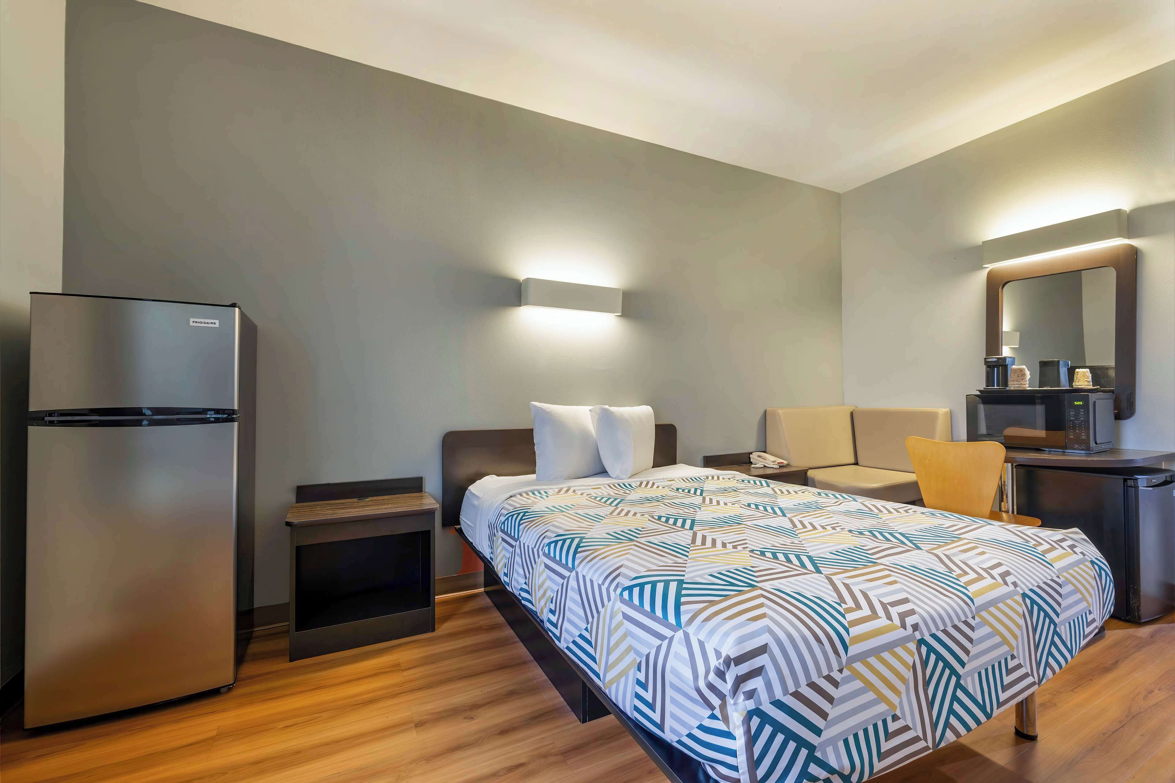 A hotel room with a bed, a refrigerator, a desk with a chair, a microwave, and a mirror.