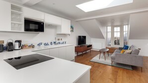 A modern kitchen with white cabinetry, a black induction cooktop, and a built-in microwave. There is a dining area with a grey sofa and a TV. - Beachfront Penthouse by the Old Course (The Scores St Andrews)