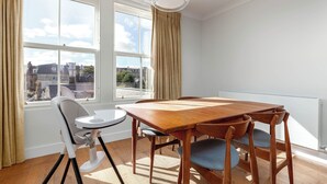 A dining area with a wooden table and chairs, a high chair, and a view of buildings through large windows. - Beachfront Penthouse by the Old Course (The Scores St Andrews)