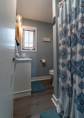 Ein Badezimmer mit Toilette, Handtuchhalter, einem Fenster mit Jalousien, einem Duschvorhang mit geometrischem Muster und einem kleinen Waschbecken mit einem darüberhängenden Handtuch.