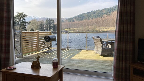Una habitación con un amplio ventanal corredera que da a una terraza exterior con vistas a un lago y montañas.