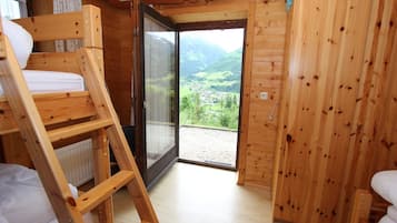 Une cabane en bois avec un lit superposé, une porte-fenêtre coulissante donnant sur un balcon avec vue sur une vallée, et une armoire en bois.